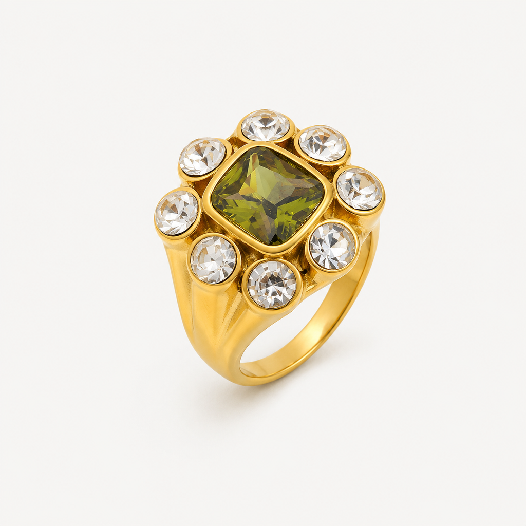 Bague Magnolia dorée à large anneau avec pierre carrée verte centrale entourée de huit pierres rondes transparentes sur fond clair