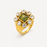 Bague Magnolia dorée à large anneau avec pierre carrée verte centrale entourée de huit pierres rondes transparentes sur fond clair
