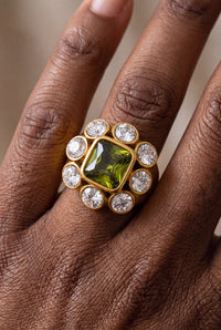 Bague Magnolia dorée portée à l'annulaire, avec pierre carrée verte entourée de huit pierres rondes transparentes